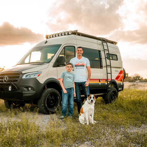 Steve, Luke, and Pickles the dog standing in front of an Adventure Van in a field of green grass at sunset
