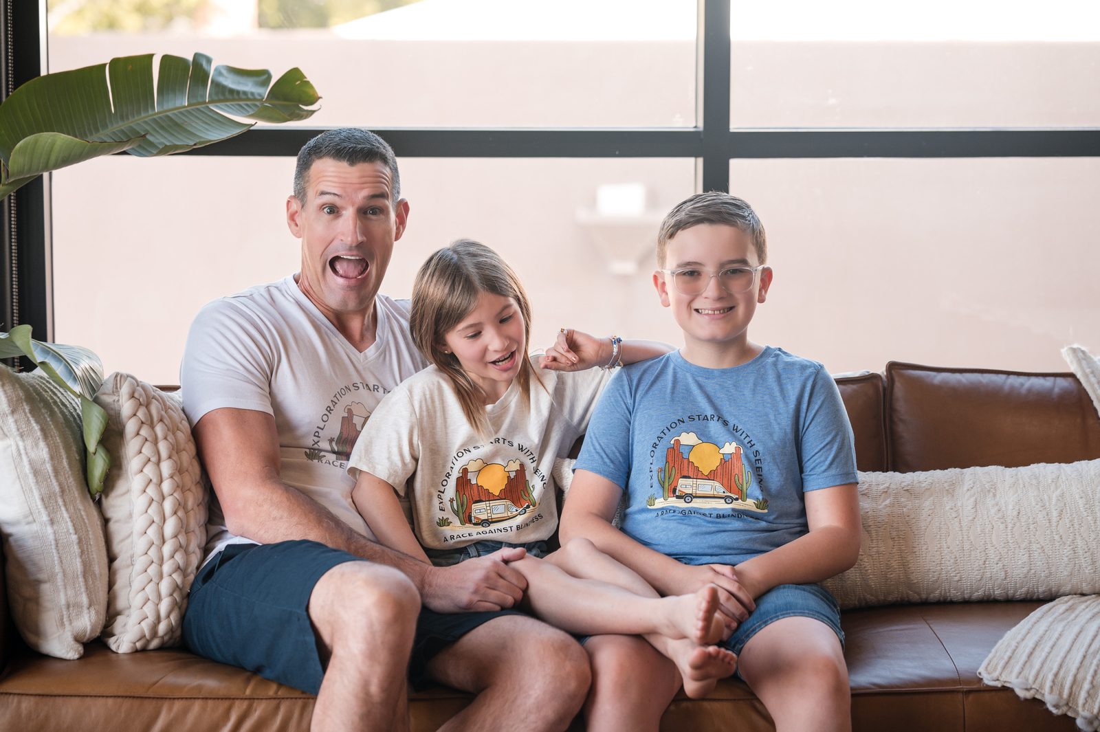 A father and two children laugh together on a living-room couch, wearing matching RAB shirts.