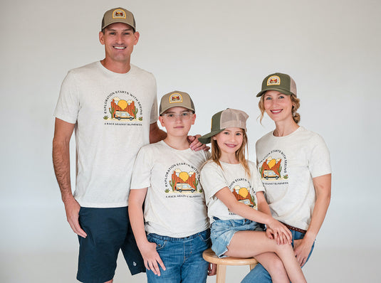 The Johnston family stands smiling proudly in front of a white background all wearing A Race Against Blindness swag shirts and hats. From left to right: Stephen, Luke, Tyler, and Kristina.