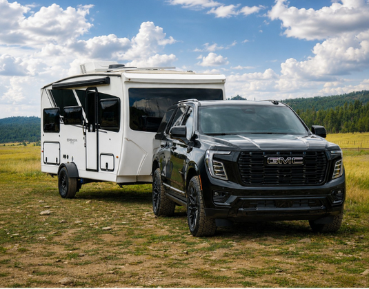 A black GMC pickup truck towing a white motorhome trailer in an open grassy field under a blue sky with white clouds.